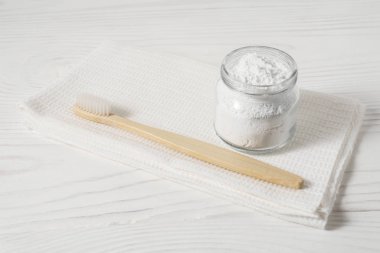 Bamboo toothbrush with a white textile towel and powder for cleaning teeth in a bowl on a light wooden background.