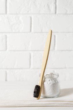 Bamboo toothbrush with a white towel and tooth powder in a bowl on a background of a light brick wall.