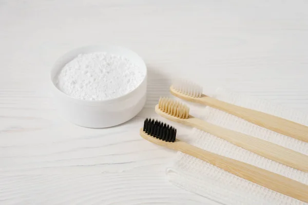 Three bamboo toothbrushes with a white textile towel and powder for brushing teeth in a bowl on a light wooden background. — Stock Fotó