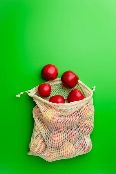 Grocery cotton net bag with red apples on a green background. Flat lay, copy space.