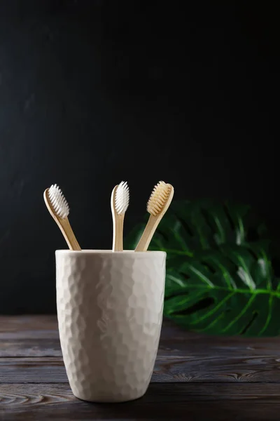 White ceramic glass with three biodegradable bamboo toothbrushes stands on a wooden table, dark background. — Stock Photo, Image