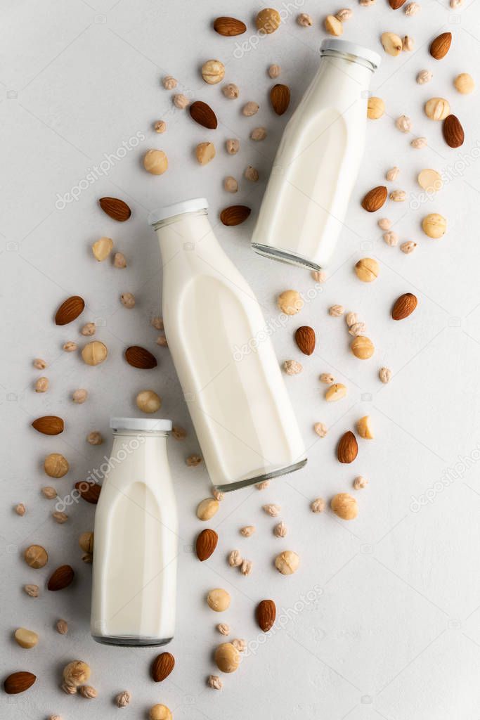 Three glass bottles with milk and scattered macadamia nuts, almonds, chickpeas on a light gray concrete surface.