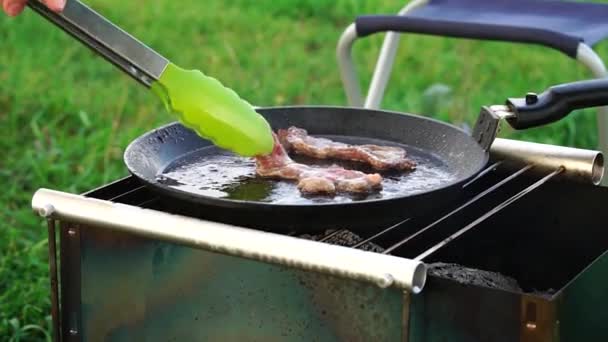 Frying bacon in a pan over charcoal during a picnic in nature. — 비디오