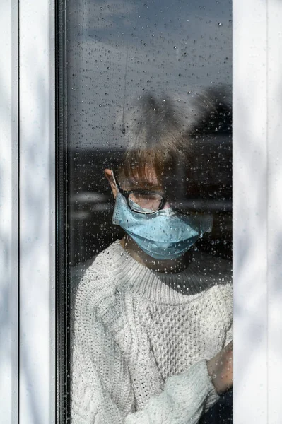 Pandemia di coronavirus 2020. Quarantena contro l'infezione e diffusione del concetto di virus COVID-19. Ragazza adolescente in una maschera protettiva medica fuori dalla finestra in isolamento domestico . — Foto Stock