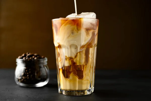 Mælk hældes i et gennemsigtigt glas med iskaffe på et sort bord . - Stock-foto