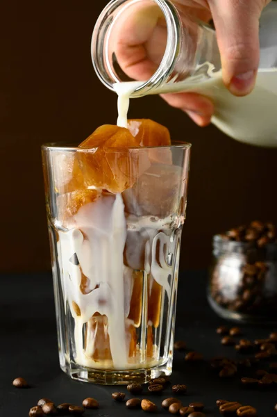 Męska ręka wlewa mleko z butelki do szklanki lodu. Mrożona kawa na ciemnym stole, brązowe tło. — Zdjęcie stockowe