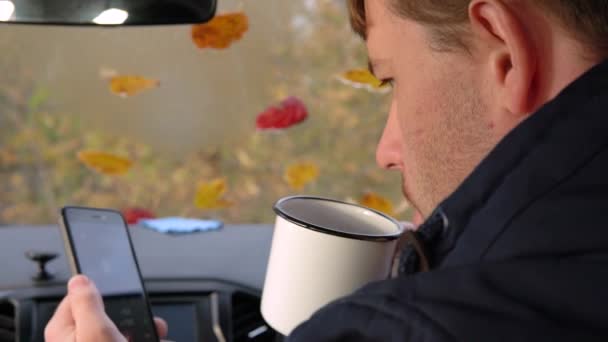 Um homem adulto senta-se dentro de um carro, bebe chá de uma caneca, usa um smartphone . — Vídeo de Stock