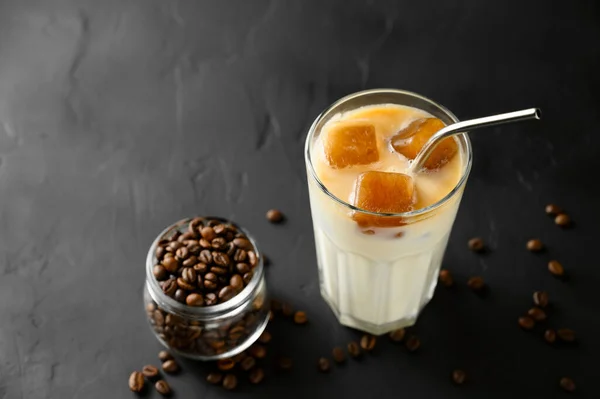 Transparent glass with milk and ice cubes made from coffee. Iced coffee with a metal tube and grains in a jar on a dark background. — Stock Photo, Image