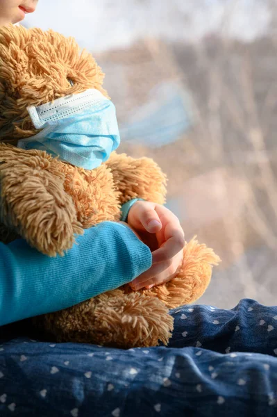Une fille dans un pull bleu s'assoit sur un rebord de fenêtre et tient un ours en peluche dans un masque de protection médicale . — Photo