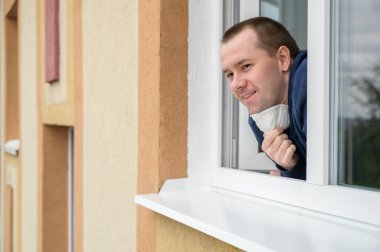 Kafkasyalı yetişkin adam gülümser, apartman penceresinden dışarı bakar ve koruyucu maskeyi çıkarır..