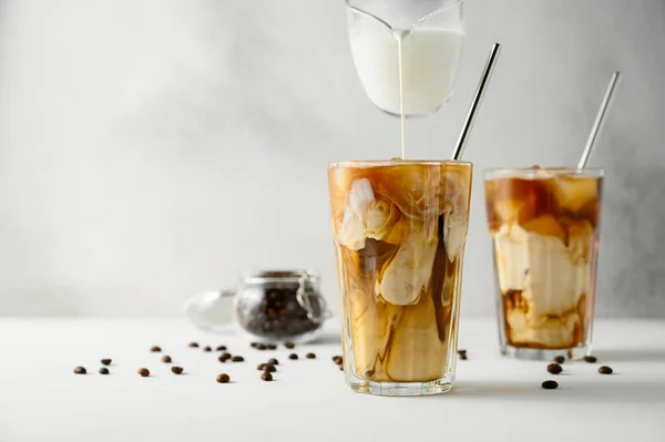 Mleko wlewa się do kawy z lodem na jasnym tle. Dwie przezroczyste szklanki orzeźwiającej mrożonej kawy na białym stole. — Zdjęcie stockowe