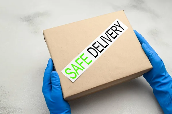 Safe delivery of packages at home during the coronavirus pandemic. Couriers hands in blue medical gloves are holding a cardboard box on a light background.