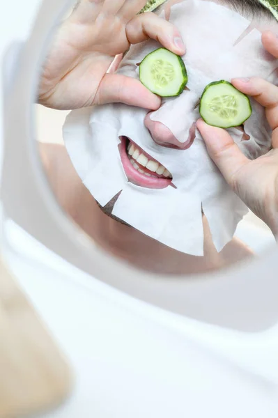 Caucasian cheerful man with a cosmetic mask on his face, holds slices of cucumber in front of his eyes. Home spa, facial skin care. — Stock Photo, Image