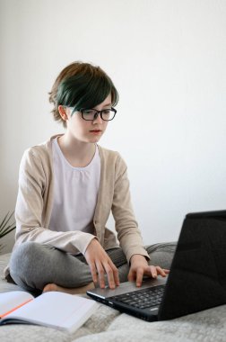 Uzaktan öğrenme çevrimiçi eğitim. Evde ders çalışan genç bir kız, dizüstü bilgisayar kullanıyor. Beyaz kız yatakta oturuyor ve okul ödevlerini yapıyor..