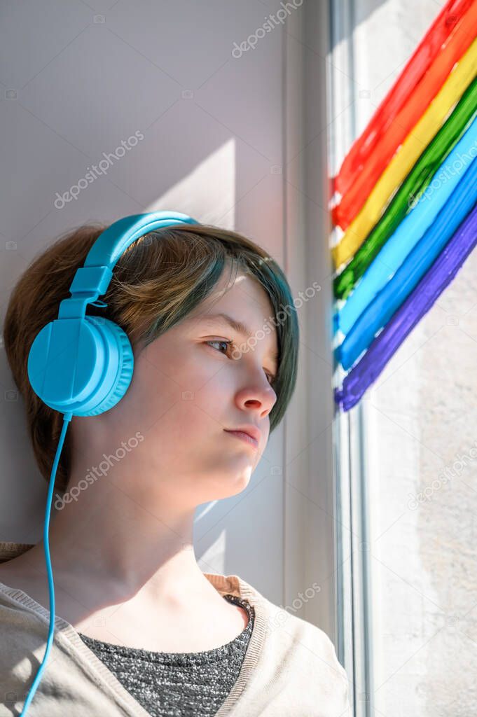 Sad teenage girl in headphones listening to music, leaning on the window. Misses her friends.