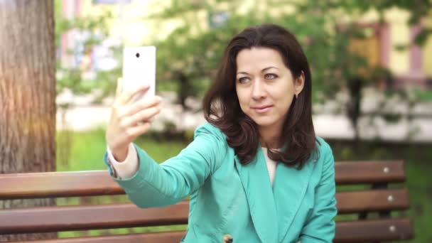 Ευτυχισμένη γυναίκα λαμβάνοντας μια selfie σε ένα παγκάκι στο πάρκο — Αρχείο Βίντεο