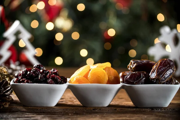 Getrocknete Aprikosen, entsteinte und getrocknete Beeren in einer Schüssel auf dem Brett mit unscharfem Weihnachtshintergrund. — Stockfoto