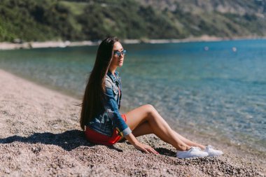 sahilde oturup güneş gözlüklü genç kadın.