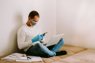 Evde karantina sırasında dizüstü bilgisayarı ve akıllı telefonu olan bir adam. SELF-ISOLATION adamı kavramı Coronavirus COVID-19 sırasında evde çalışıyor..