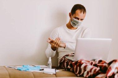 Adam evde virüs önleme için sıvı el dezenfektanı kullanıyor. Evde dizüstü bilgisayar başında çalışan ve elleri dezenfekte eden bir adam. Evde kal.