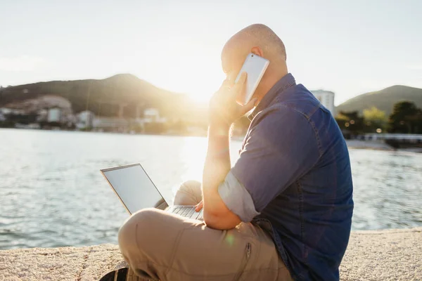 man making online reservation using phone and laptop on beach. man talking over phone and using laptop at seaside beach at sunset. Man working remotely online using laptop sitting on beach at sunset time.