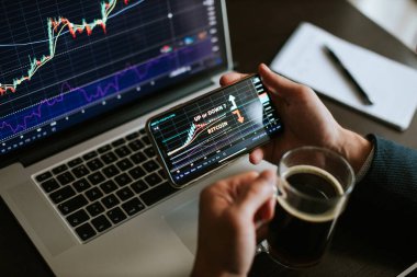 Yatırım borsacısı dizüstü bilgisayar ve telefon kullanarak bitcoin fiyat hareketini tahmin ediyor. Kristal tüccar finansal yöneticisi hisse senedi grafiğini analiz ediyor. Yönetici birden fazla aygıt ve kahve kullanıyor..