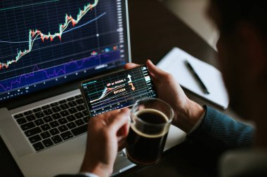 Telefon ve dizüstü bilgisayar kullanarak bitcoin fiyat artışını ya da düşüş eğilimini analiz eden borsacı. Kripto para birimi tüccarı birden fazla cihaz kullanarak fiyat hareketini tahmin ediyor ve kahve içiyor..