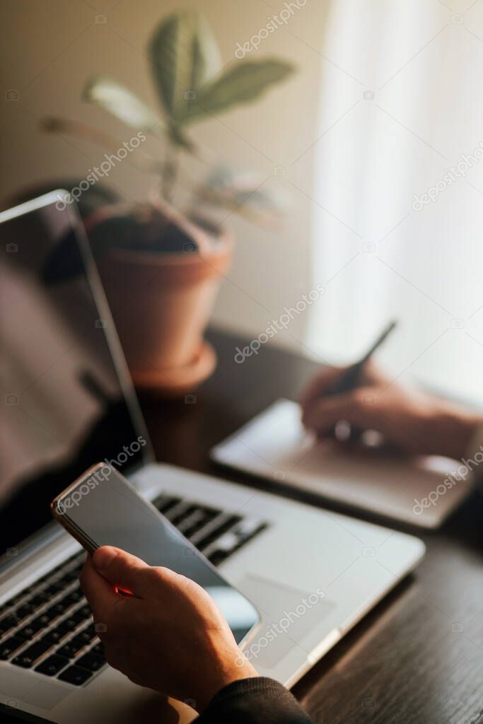 office worker browsing phone and laptop indoors. businessman using phone and laptop in office workspace with plant background. Black screen gadgets. Handwriting notes during work day