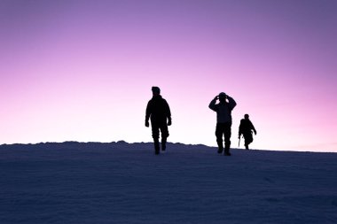 Üç adam gün batımına tırmandıktan sonra tepeden dağlara iniyor. Pembe, açık gökyüzü, kış.