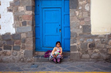 Peru, Cusco 2019-11-29. Milli giysili küçük bir kız kapının önünde bebek bir lama ile oturuyor. Çocukluktan beri çalışmaya zorlanan çocukların zor hayatı kavramı.