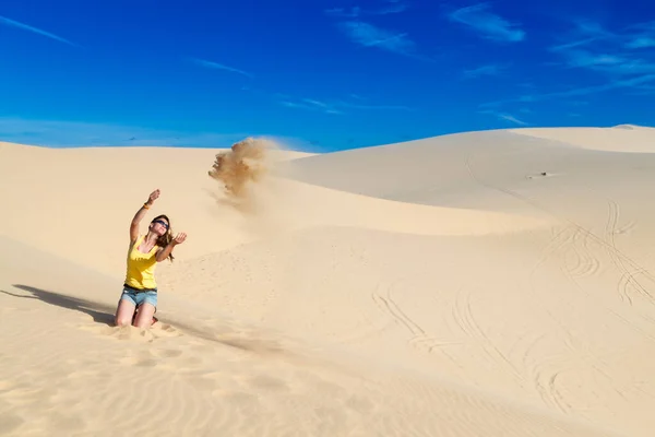 Woman sits on the sand in the desert, throws sand into the air, blue sky