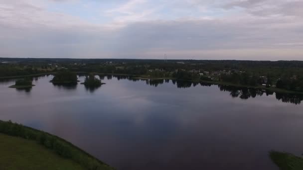 Una vista desde el aire a un pequeño lago tranquilo. Verano, costas verdes, nubes se reflejan. En la orilla lejana hay pequeñas casas — Vídeo de stock