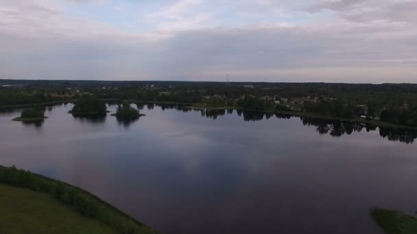 En vy från luften till en liten lugn sjö. Sommar, gröna stränder, moln återspeglas. På den bortersta stranden är små hus — Stockvideo