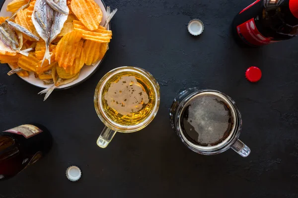 Becher mit Bier, Flaschen und Snacks auf schwarzem Hintergrund — Stockfoto