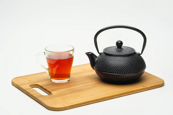 Чугунный чайник на деревянной доске и стакан с горячим чаем на белом фоне . — стоковое фото