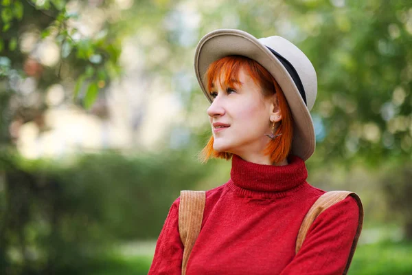 Kopfporträt eines jungen attraktiven Mädchens mit roten Haaren, Hut und rotem Pullover vor einem Hintergrund grüner Bäume im Park. — Stockfoto