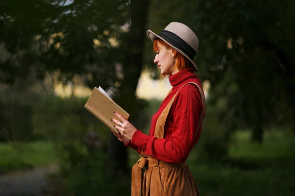 Retrato en la cabeza de una joven atractiva con el pelo rojo en sombrero y suéter rojo sobre el fondo de árboles verdes en el parque. Ella sostiene el libro en las manos y lee . —  Fotos de Stock