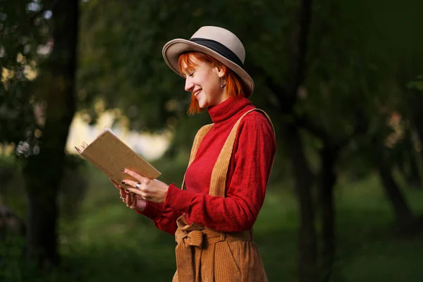 Retrato en la cabeza de una joven atractiva con el pelo rojo en sombrero y suéter rojo sobre el fondo de árboles verdes en el parque. Ella sostiene el libro en las manos y lee . —  Fotos de Stock