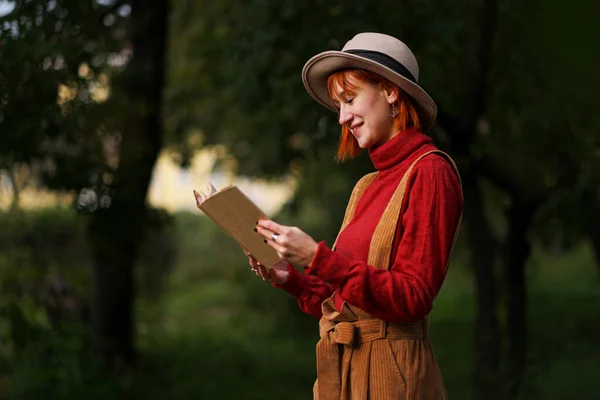 Portrait en tête d'affiche d'une jeune fille séduisante aux cheveux roux en chapeau et pull rouge sur fond d'arbres verts dans le parc. Elle tient le livre dans ses mains et lit : . — Photo