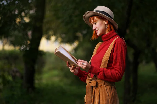 Retrato en la cabeza de una joven atractiva con el pelo rojo en sombrero y suéter rojo sobre el fondo de árboles verdes en el parque. Ella sostiene el libro en las manos y lee . —  Fotos de Stock