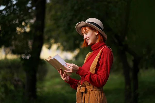 Portrait en tête d'affiche d'une jeune fille séduisante aux cheveux roux en chapeau et pull rouge sur fond d'arbres verts dans le parc. Elle tient le livre dans ses mains et lit : . — Photo