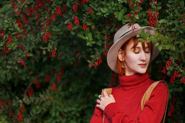 Porträt eines jungen attraktiven Mädchens mit roten Haaren, Hut und rotem Pullover vor dem Hintergrund eines Berberitzenstrauches mit roten Beeren. — Stockfoto