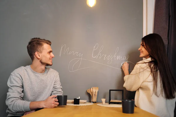 Casal jovem casal em roupas casuais, homem e mulher, sentado em casa na cozinha à mesa. Desenhe Feliz Natal com giz na parede . — Fotografia de Stock