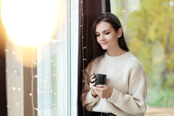 Portrét mladé krásné ženy v teplém svetru a s šálkem teplého nápoje doma na pozadí okna. Lampy teplého světla. — Stock fotografie