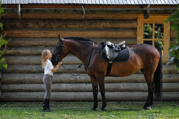 Una jinete adolescente se para junto a un caballo marrón y la abraza. Sobre el fondo de un establo de madera . — Foto de Stock