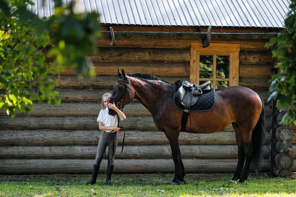 Жокей-подросток стоит рядом с коричневой лошадью и обнимает её. На фоне деревянных настенных конюшен . — стоковое фото