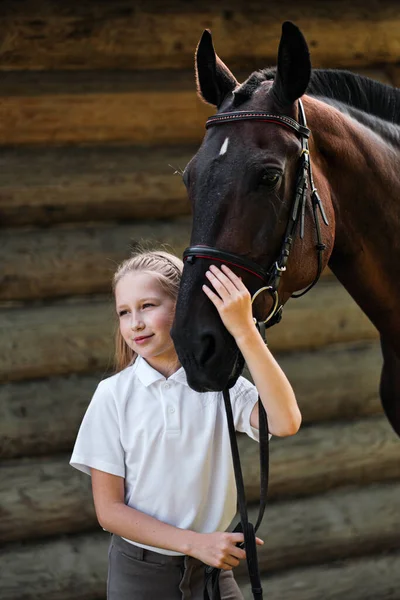 Una jinete adolescente se para junto a un caballo marrón y la abraza. Sobre el fondo de un establo de madera . — Foto de Stock
