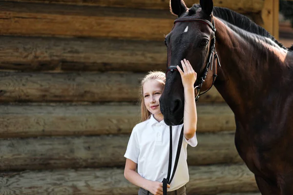 A teenage girl jockey stands next to a brown horse and hugs her. Against the background of a wooden wall stables.