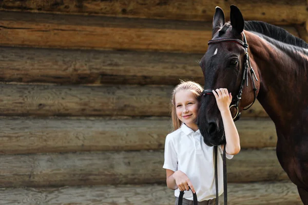 Una jinete adolescente se para junto a un caballo marrón y la abraza. Sobre el fondo de un establo de madera . — Foto de Stock