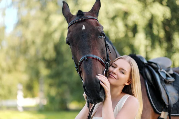 Joven mujer rubia atractiva abraza a un caballo marrón . — Foto de Stock
